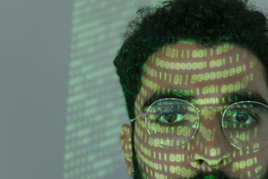 A close-up of a young man with short, curly dark hair and round glasses with clear lenses, reflecting green digital binary code across his face. The background shows a blurred wall of horizontal green