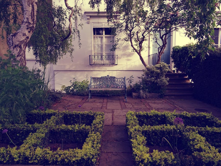 A small, neatly maintained garden area in front of a white building at Golders Green station, featuring a paved pathway leading to a decorative metal bench. The garden is bordered by low, trimmed hedg